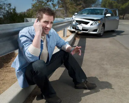 A man sitting on a curb talking on his phone after a car accident, with the damaged car behind him